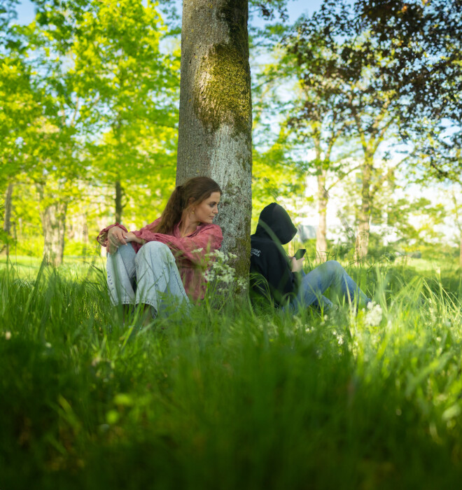Begeleider jeugdzorg met open houding en een jongere met gesloten houding zitten tegen een boom.
